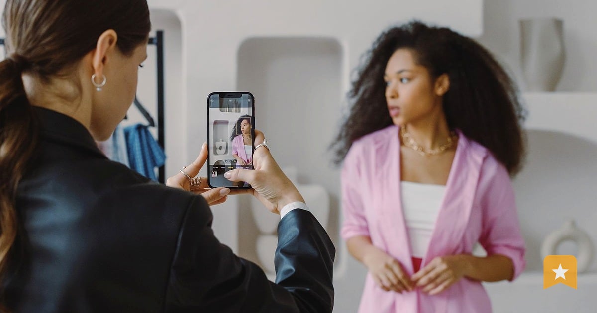 Woman in pink shirt being photographed by another woman holding a phone, bright white interior with shelving behind them