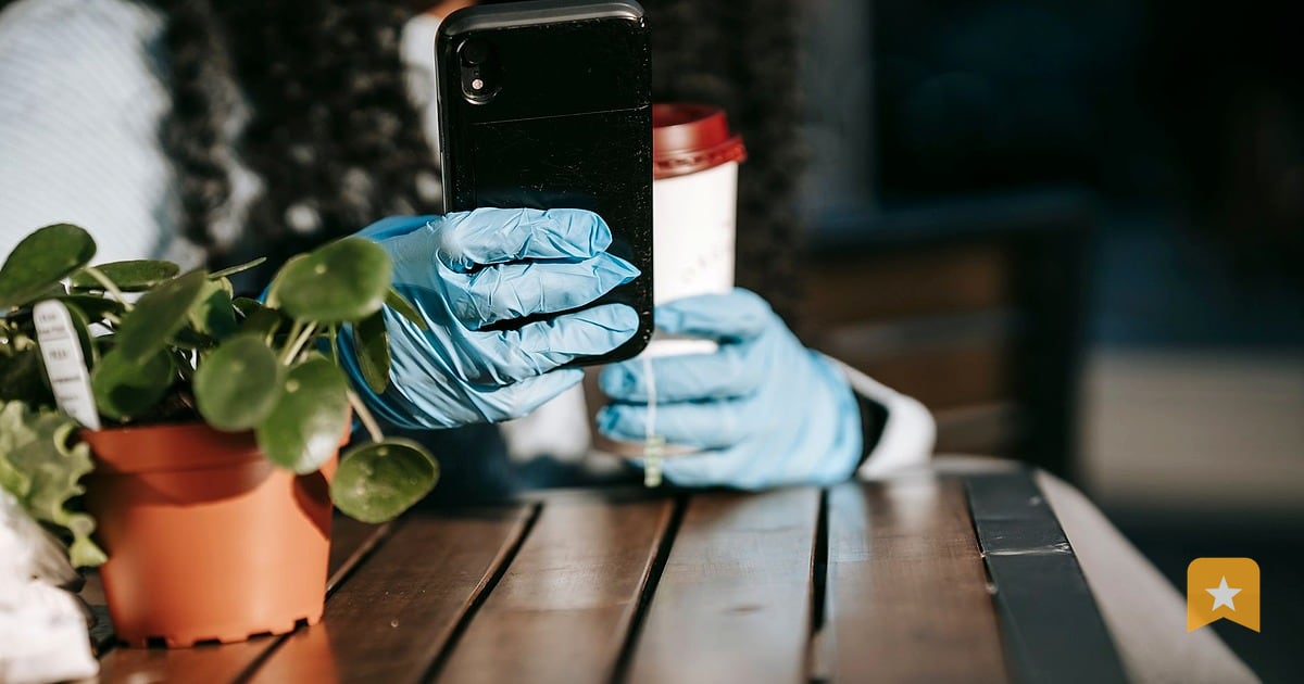 Person in blue gloves holding phone at outdoor cafe table with potted plant and beverage