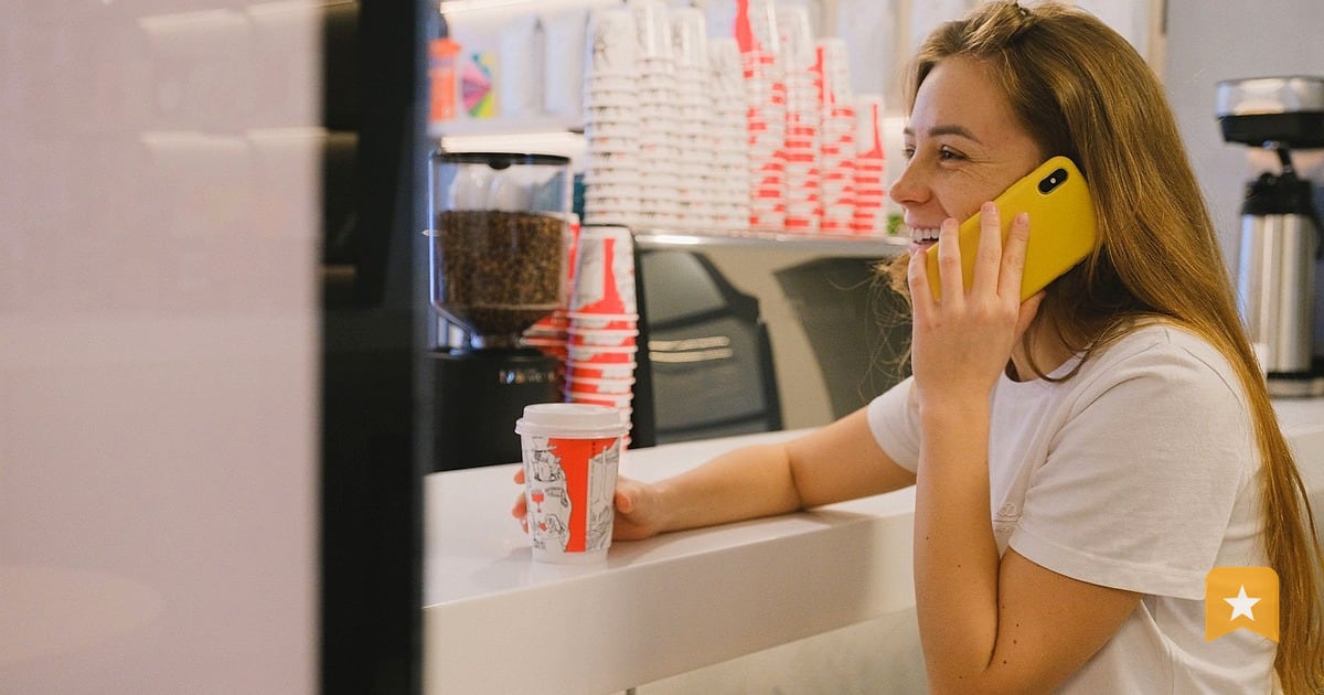 Woman in white shirt holding yellow phone at cafe counter with drink, engaged in conversation; owner checking customer feedback.
