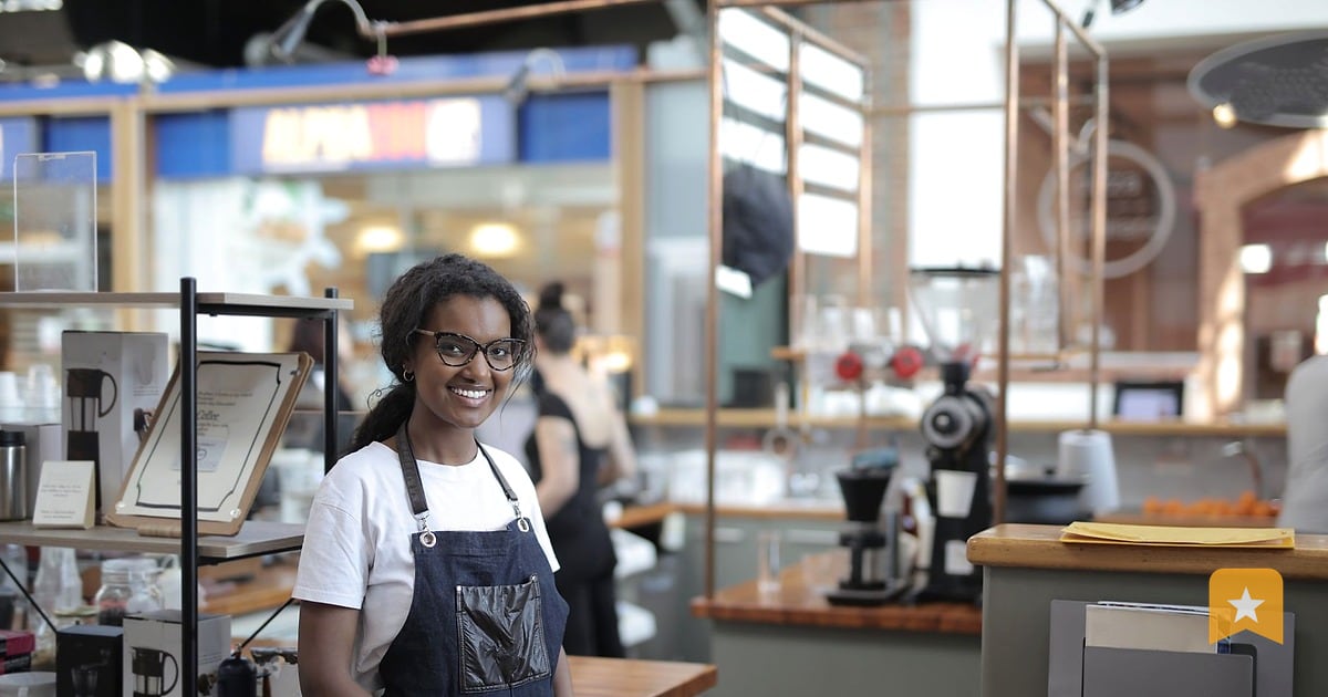 Small business owner in apron standing behind counter in bright, modern cafe with espresso machine and customers in background