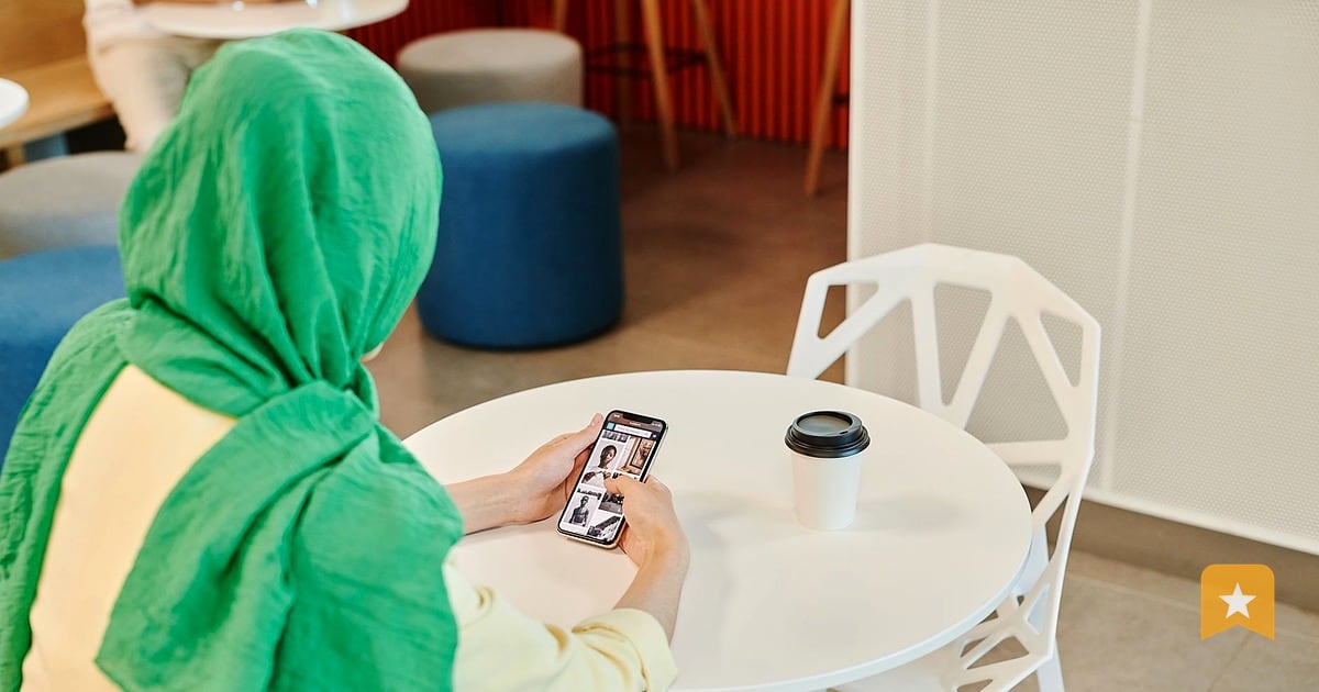 Person in green scarf holding phone at white cafe table, reviewing or responding to online feedback from a customer.