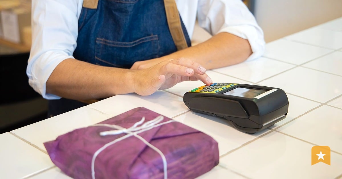 Small business owner in apron standing at counter with payment terminal, ready to process a customer transaction at her shop.
