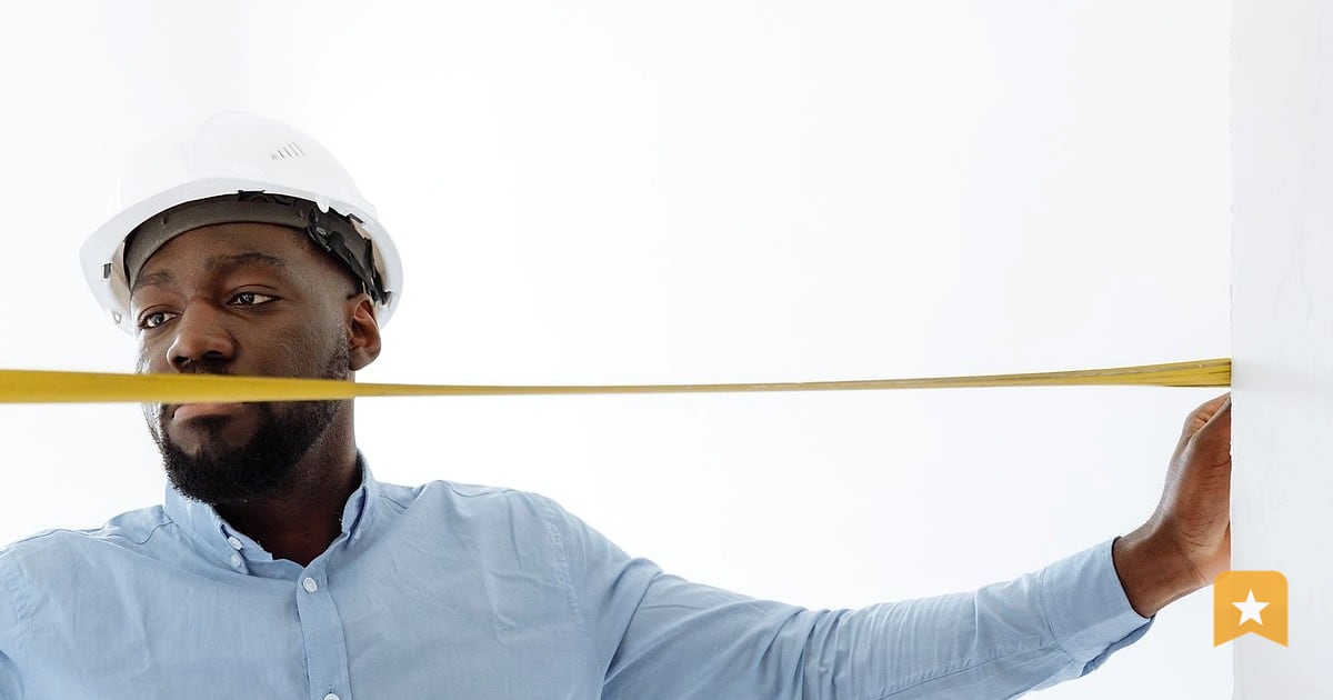 Contractor in hard hat and blue shirt holding a measuring tape across a white wall indoors