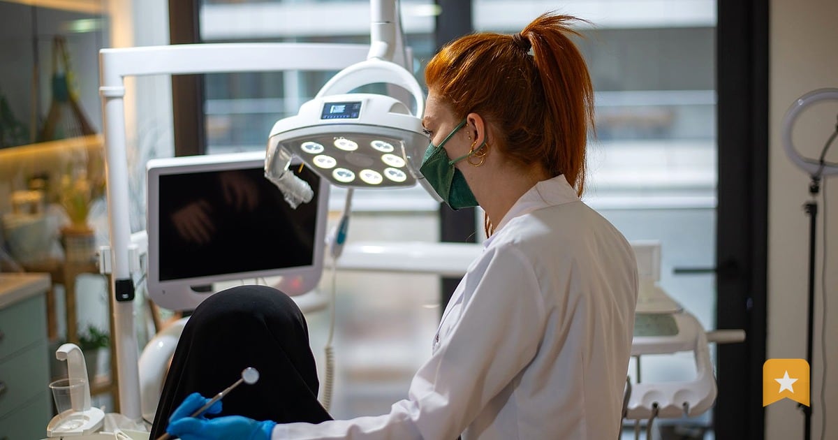 Dentist in white coat and blue gloves works with patient in modern clinic chair, overhead surgical light visible