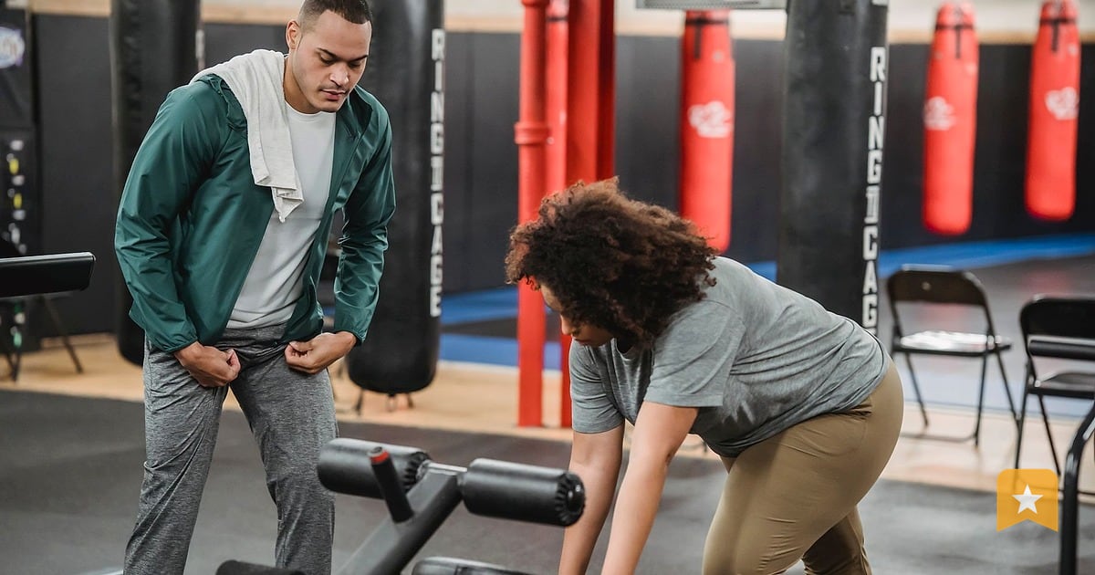Gym trainer watches client perform an exercise on equipment in a well-lit studio with punching bags and weights visible