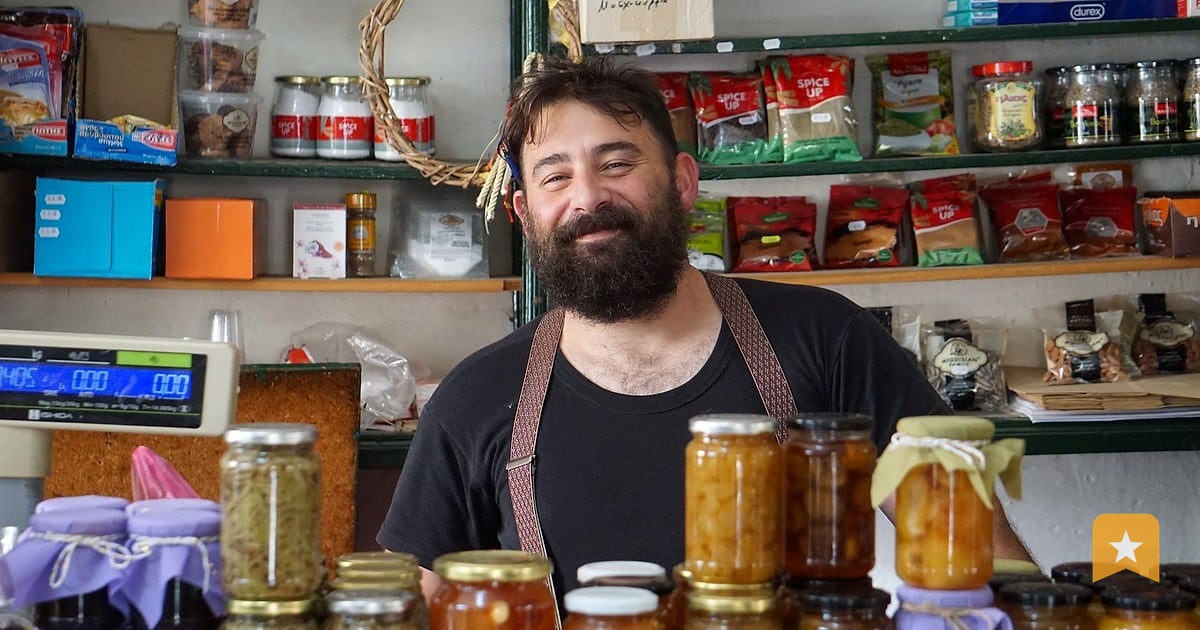 Small business owner with beard and apron standing behind counter lined with glass jars of preserves